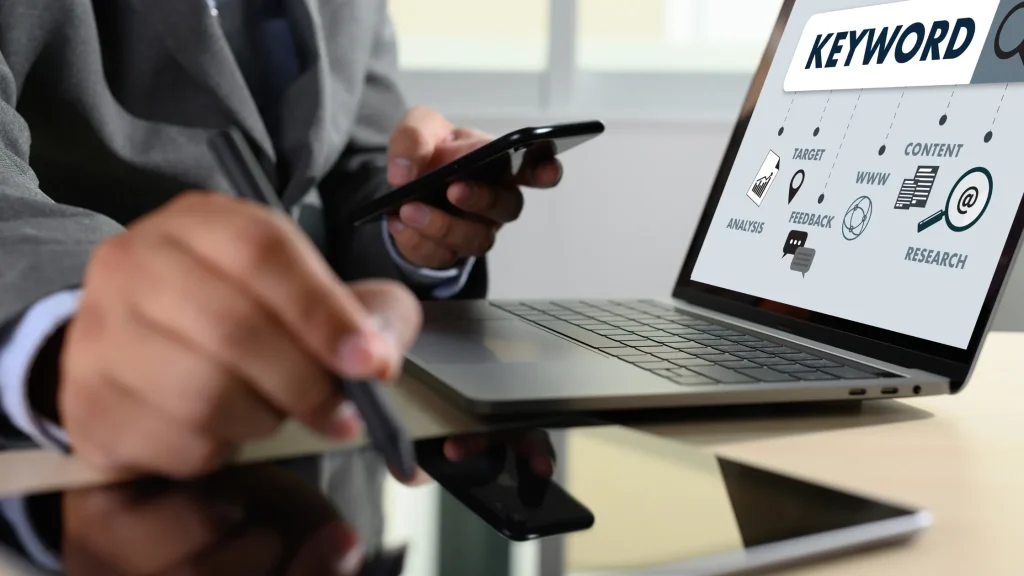 A business professional using a laptop and smartphone, analyzing keyword research, content strategy, and SEO elements displayed on the screen.